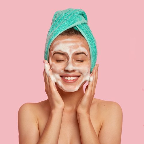 pleased-smiling-woman-washes-face-with-cleansing-gel-has-soap-on-complexion-keeps-eyes-shut-wears-wrapped-towel-on-head-has-naked-body