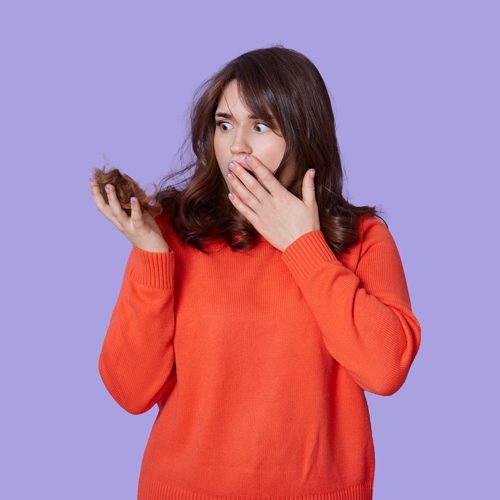 shocked-female-having-problems-with-hair-loss-holding-curl-and-covering-mouth-with-palm-lady-wearing-casual-orange-jumper-being-frustrated-of-balding-problems-isolated-over-blue-wall